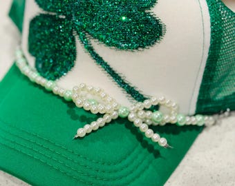 Puede incluir: Un gorro de camionero blanco y verde con un trébol verde brillante de gran tamaño y un ribete de perlas. El gorro tiene una malla verde en la parte posterior.