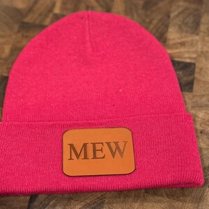 May include: A pink beanie with a brown leather patch that says "MEW" on it.