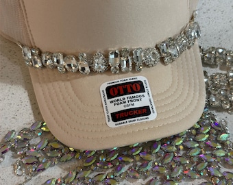 Könnte beinhalten: Eine beige Trucker-Cap mit einer Reihe klarer, facettierter Strasssteine auf der Vorderseite. Ein rechteckiger Aufkleber auf dem Schirm trägt die Aufschrift OTTO WORLD FAMOUS FOAM FRONT OSFM TRUCKER. Darunter liegen weitere Stränge aus irisierenden Strasssteinen.