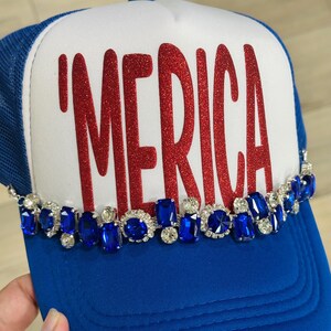 Op de afbeelding: Een blauw-witte truckerpet met de rode glittertekst 'MERICA op het voorpaneel. De onderkant van het voorpaneel is versierd met een rij afwisselend blauwe en doorzichtige gefacetteerde strass-steentjes in zilverkleurige zettingen.