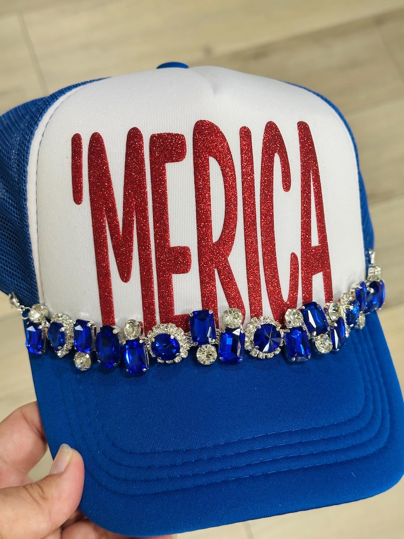 Op de afbeelding: Een blauw-witte truckerpet met de rode glittertekst 'MERICA op het voorpaneel. De onderkant van het voorpaneel is versierd met een rij afwisselend blauwe en doorzichtige gefacetteerde strass-steentjes in zilverkleurige zettingen.