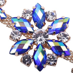 K&ouml;nnte beinhalten: Nahaufnahme eines goldfarbenen Schmuckst&uuml;cks mit floralem Design. Das Schmuckst&uuml;ck ist mit leuchtend blauen und klaren, kristallartigen Steinen besetzt. Die Steine sind in Bl&uuml;tenform angeordnet, mit einem zentralen klaren Stein.