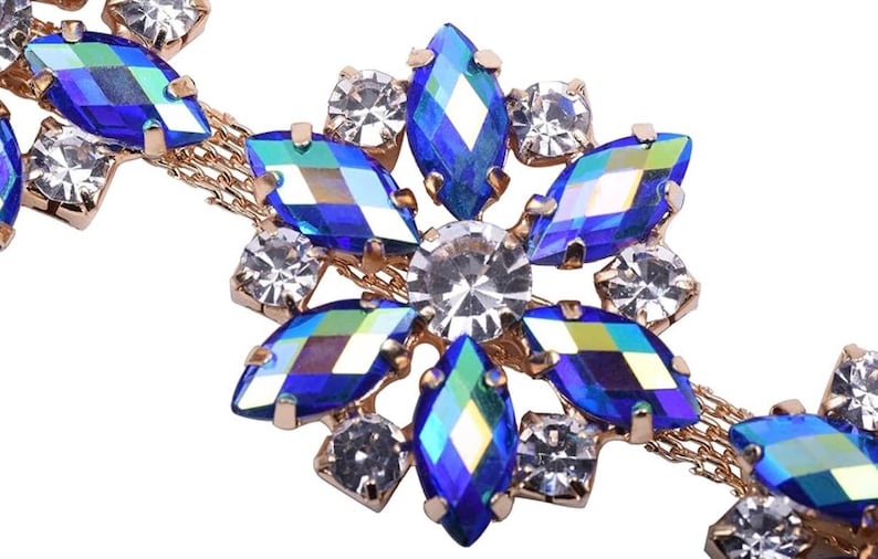 K&ouml;nnte beinhalten: Nahaufnahme eines goldfarbenen Schmuckst&uuml;cks mit floralem Design. Das Schmuckst&uuml;ck ist mit leuchtend blauen und klaren, kristallartigen Steinen besetzt. Die Steine sind in Bl&uuml;tenform angeordnet, mit einem zentralen klaren Stein.