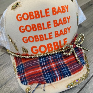 May include: A cream-colored trucker hat with the words "GOBBLE BABY" in orange. The brim features a red, white, and blue plaid pattern, gold beads, and glitter accents. A decorative bow is attached.