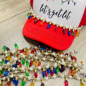 May include: Red and white trucker hat with the text "let's get lit" and a string of colorful Christmas lights. The hat is decorated with a string of faux pearls and colorful light bulbs.