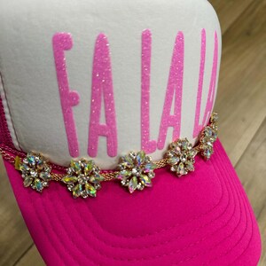 K&ouml;nnte beinhalten: Eine pinkfarbene Trucker-M&uuml;tze mit wei&szlig;em Frontpanel. Auf der M&uuml;tze steht das Wort "FA LA LA" in glitzernden pinkfarbenen Buchstaben. Eine goldene Kette mit dekorativen floralen Akzenten verziert den Rand und sorgt f&uuml;r einen Hauch von Glanz und Glamour.