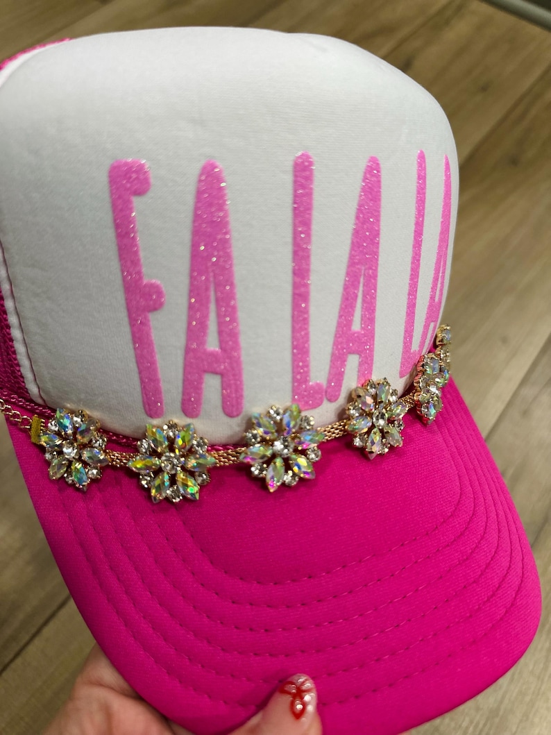 K&ouml;nnte beinhalten: Eine pinkfarbene Trucker-M&uuml;tze mit wei&szlig;em Frontpanel. Auf der M&uuml;tze steht das Wort "FA LA LA" in glitzernden pinkfarbenen Buchstaben. Eine goldene Kette mit dekorativen floralen Akzenten verziert den Rand und sorgt f&uuml;r einen Hauch von Glanz und Glamour.