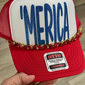 Op de afbeelding: Een rood-witte truckerpet met de tekst 'MERICA in blauwe glitterletters op het voorpaneel van schuim. De pet is versierd met een decoratieve ketting van rode strass-steentjes aan de onderkant van het voorpaneel en bevat een Otto-sticker.