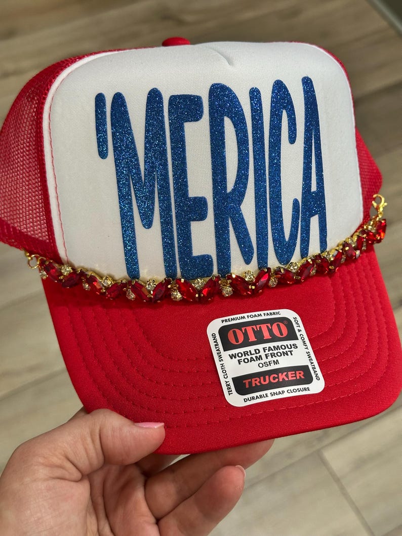 Op de afbeelding: Een rood-witte truckerpet met de tekst 'MERICA in blauwe glitterletters op het voorpaneel van schuim. De pet is versierd met een decoratieve ketting van rode strass-steentjes aan de onderkant van het voorpaneel en bevat een Otto-sticker.