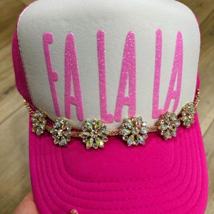 K&ouml;nnte beinhalten: Eine leuchtend pinkfarbene Trucker-M&uuml;tze mit wei&szlig;er Front. Der Text "FA LA LA" ist in glitzerndem Pink aufgedruckt. Eine goldene Kette mit floralen Strass-Akzenten verziert den Schirm.