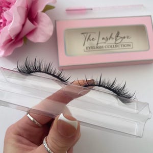 May include: A close-up of a pair of false eyelashes on a clear plastic tray. The eyelashes are black and have a natural, wispy look. The tray is labeled "The Lashbox Eyelash Collection."