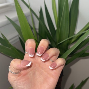 Könnte beinhalten: Nahaufnahme einer Hand mit kurzen, quadratischen Nägeln, die mit einer hellrosa Basis und weißen Spitzen lackiert sind. Die Nägel haben einen French-Manicure-Stil.
