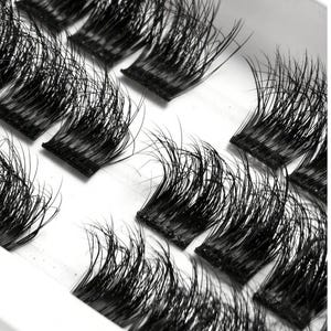 Pode incluir: Close-up de vários conjuntos de cílios postiços pretos. Cada conjunto apresenta uma faixa fina e flexível com inúmeros fios de cílios individuais, criando uma aparência completa e volumosa. Os cílios estão dispostos em um recipiente branco, prontos para aplicação.