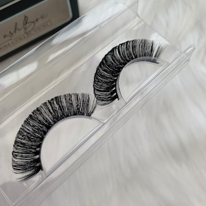 May include: Two pairs of black false eyelashes in a clear plastic container. The eyelashes are long and full, with a natural curl.