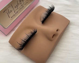 May include: A pair of false eyelashes displayed on a brown mannequin head. The eyelashes are long and thick, and they are arranged in a natural-looking way. The mannequin head is made of a soft, pliable material, and it is designed to hold the eyelashes in place. False eyelashes are a popular beauty product that can be used to enhance the appearance of the eyes.