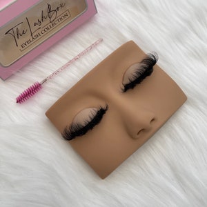 May include: A tan mannequin head with closed eyes and black eyelashes. A pink-handled lash brush and a pink box with the text "The Lash Box EYELASH COLLECTION" are also visible.