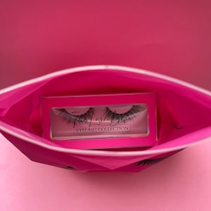 Könnte beinhalten: Eine rosa Reißverschlusstasche mit einem schwarzen und weißen Wimpern-Design. Die Tasche ist geöffnet und eine durchsichtige Plastikbox mit einem Paar schwarzer künstlicher Wimpern befindet sich im Inneren. Auf der Box steht "The Lash Box Eyelash Collection".