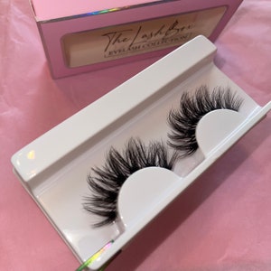 Könnte beinhalten: Zwei Paar schwarze falsche Wimpern in einem weißen Plastikbehälter. Der Behälter trägt den Text "The Lash Box Eyelash Collection".