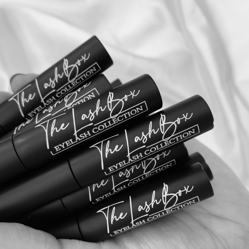 May include: A stack of black tubes with white lettering that reads "The Lash Box Eyelash Collection".