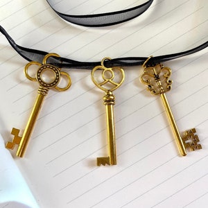 May include: Three gold-toned skeleton keys on a black ribbon. The keys have ornate designs and are hanging on a white lined notebook.