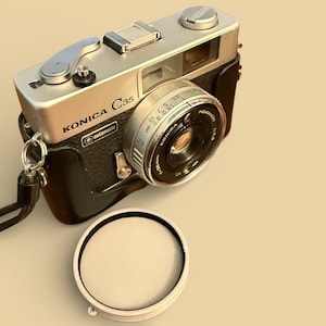 May include: A vintage Konica C35 camera with a silver top, black leather body, and a lens with numerical markings. The camera lens cap is off and placed in front of the camera. The camera has a strap attached and the word "automatic" is visible.