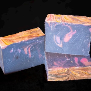 May include: Three handmade soap bars with a marbled design. The soap has a blue base with pink swirls and a top layer of yellow, orange, and brown. The bars are arranged on a black background.
