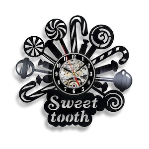 Può includere: Un orologio in vinile nero con quadrante e lancette. L'orologio è decorato con forme di bastoncini di zucchero e lecca-lecca. Il testo "Sweet tooth" è stampato sotto il quadrante.