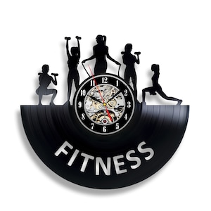 Może przedstawiać: Czarny zegar winylowy z sylwetką pięciu osób ćwiczących. Tarcza zegara ma cyfry rzymskie, a pod tarczą zegara nadrukowano napis "FITNESS".