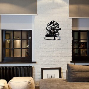 May include: A black wall clock shaped like a stack of books, with a clock face in the center. The clock is mounted on a white brick wall, between two windows with black frames. The room has a modern, minimalist design.