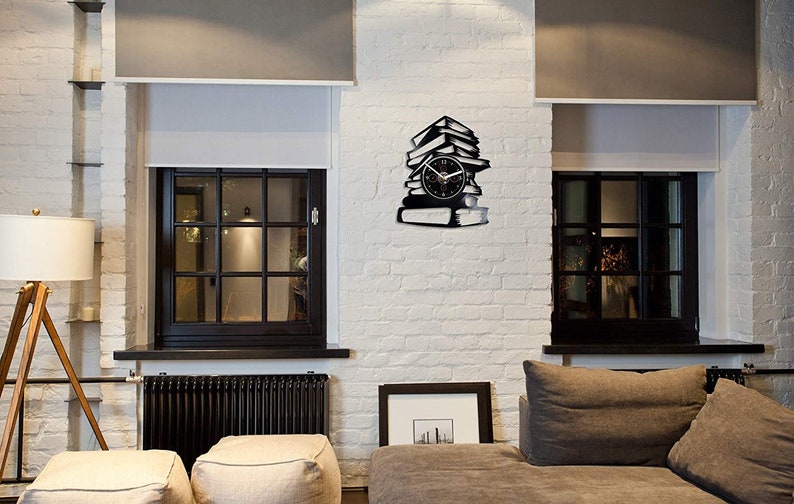 May include: A black wall clock shaped like a stack of books, with a clock face in the center. The clock is mounted on a white brick wall, between two windows with black frames. The room has a modern, minimalist design.