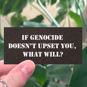 May include: A rectangular, dark brown card with the text "IF GENOCIDE DOESN'T UPSET YOU, WHAT WILL?" in white. The card is held in front of a lush green plant with large leaves.
