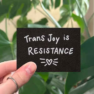 May include: A black rectangular patch with the words "Trans Joy is RESISTANCE" in white script, along with a heart symbol. The patch is held in front of a lush green plant.
