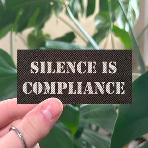 May include: A black card with white text that reads "Silence is Compliance". The card is being held by a hand in front of a green leafy plant.