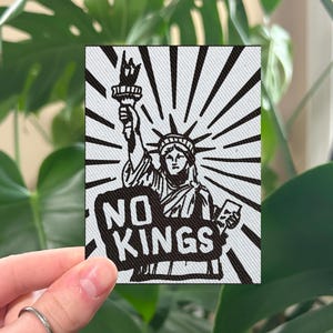 May include: A rectangular patch featuring a black and white illustration of the Statue of Liberty holding a torch, with the words "NO KINGS" in bold letters. The design is set against a backdrop of radiating lines.