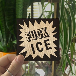 May include: Square sticker with the text "FUCK ICE" in a bold, black font, set against a beige, textured background within a black starburst design. The sticker is held in front of a backdrop of green plants.