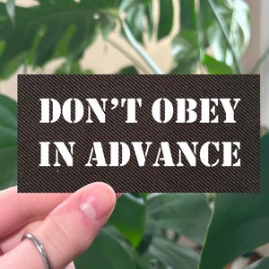 May include: A rectangular patch with the words "DON'T OBEY IN ADVANCE" in white, stencil-style lettering on a dark brown background. The patch is held up in front of a lush green plant with large leaves.