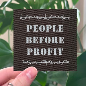 May include: A square, brown fabric patch with the words "PEOPLE BEFORE PROFIT" in white block letters. The patch is bordered by white barbed wire designs. The patch is held in front of a green leafy plant.