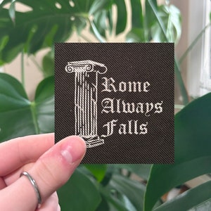 May include: A black fabric patch with white embroidery of a Roman column and the text "Rome Always Falls".