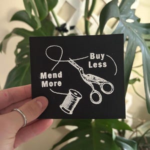 May include: Black square patch with white embroidered design. Features the words "Mend More" and "Buy Less" with a needle, thread, scissors, and spool of thread. The patch is held in front of a green leafy background.