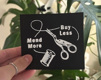May include: Black square patch with white embroidered design. Features the words "Mend More" and "Buy Less" with a needle, thread, scissors, and spool of thread. The patch is held in front of a green leafy background.
