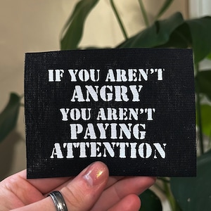 May include: Black fabric patch with white text that reads "If you aren't angry you aren't paying attention."