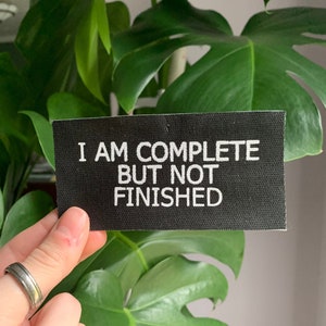 May include: Black fabric patch with white text that reads "I am complete but not finished".