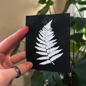 May include: A white fern frond design on a black fabric patch. The patch is made of a textured material and has a slightly rough surface.
