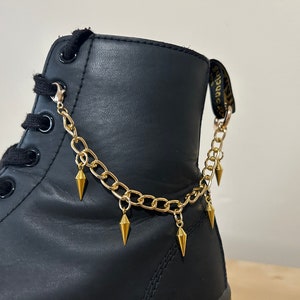 May include: Black leather boot with a gold chain and spike accents. The chain is attached to the boot with two metal clasps.