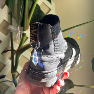 May include: Black leather ankle boots with a black elastic panel and a gold and black "AirWair" label on the back. A clear crystal charm hangs from a silver ring on the back of the boot.