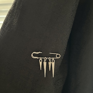 May include: A silver safety pin with five dangling silver spikes. The pin is attached to a black fabric background.