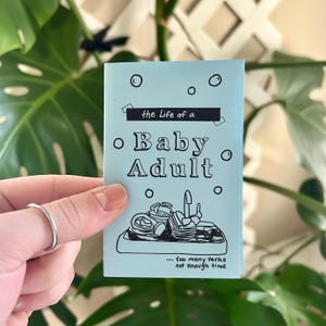 May include: A light blue booklet with the title "the Life of a Baby Adult" and a black and white illustration of a tray overflowing with dishes and utensils. The text below the illustration reads "...too many tasks not enough time."