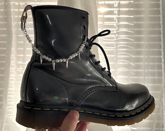 May include: Black leather boots with a silver chain and pearl accents. The boots have a thick sole and a lace-up closure.