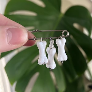 May include: A white enamel pin featuring three white bone charms hanging from a silver safety pin.