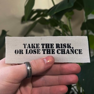May include: White fabric patch with black text that reads "Take the risk, or lose the chance."